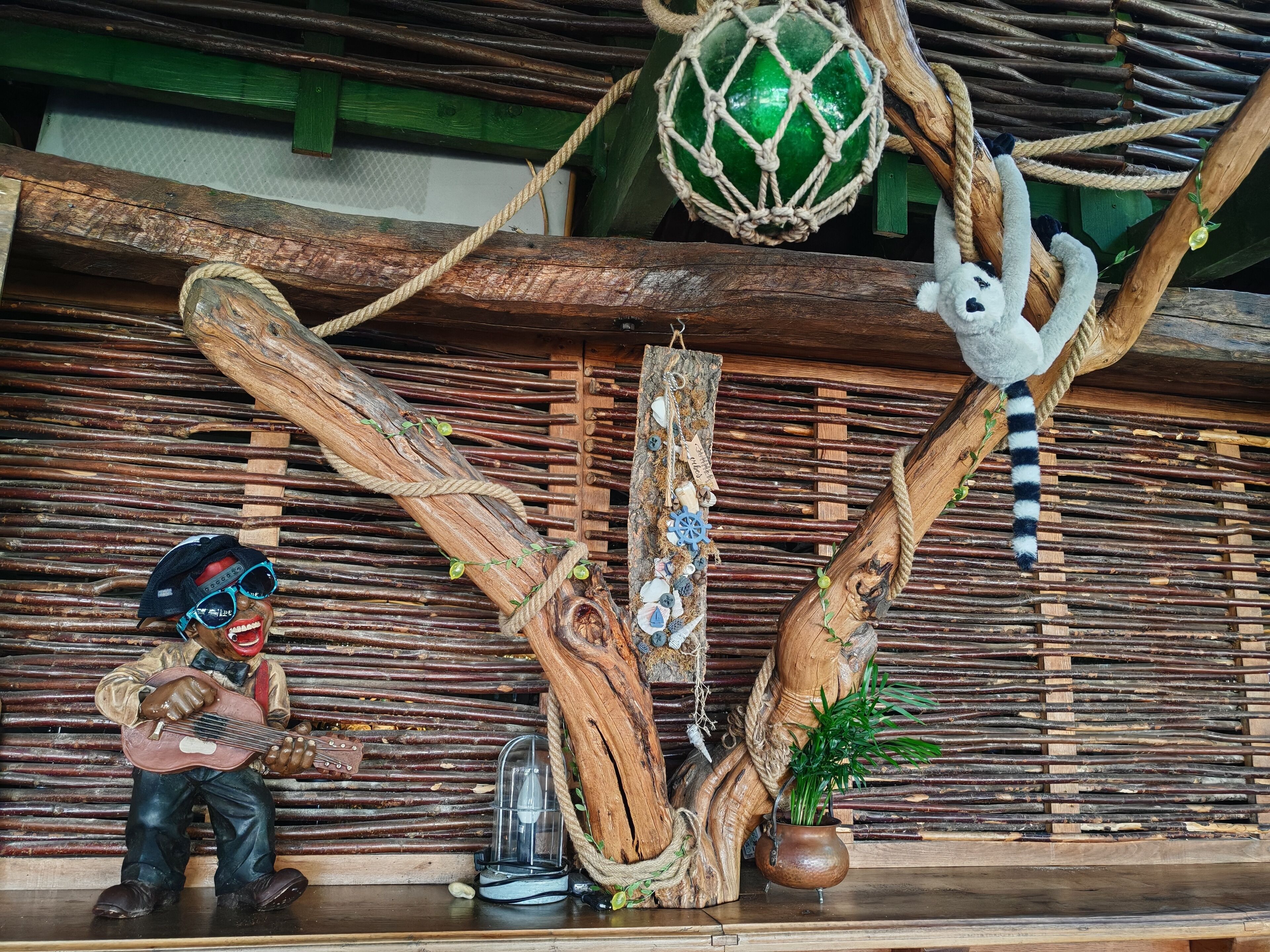 A rustic interior with a wooden structure, a hanging macrame ball, a white plush toy, a small figurine of a man playing a guitar, and a potted plant.