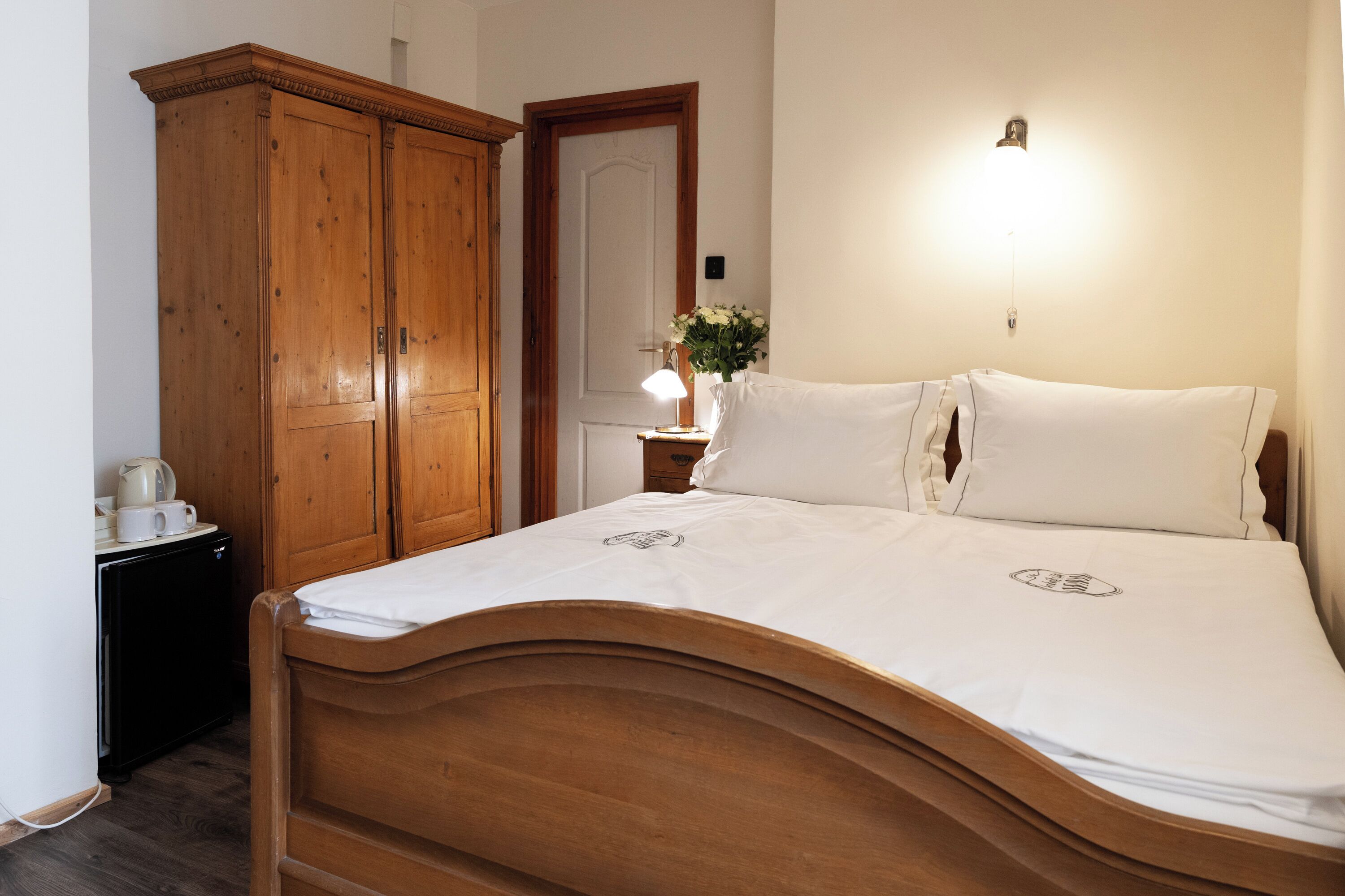 Una cama bien hecha con cabecera de madera, un armario de madera, una mesita de noche con una lámpara y un espejo en la pared.
