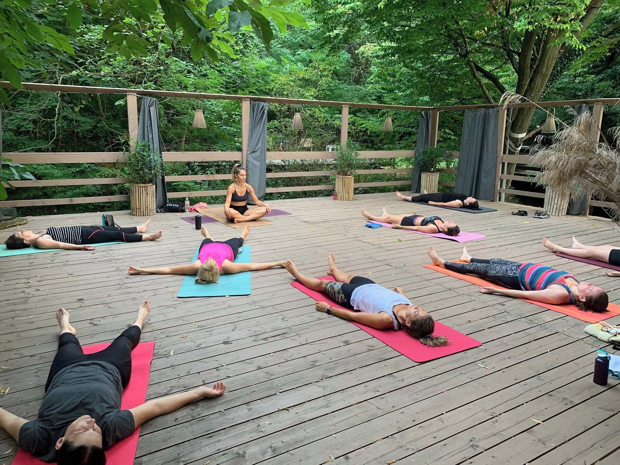 Un grupo de personas sobre esterillas de yoga en un entorno exterior con una terraza de madera y follaje de fondo.