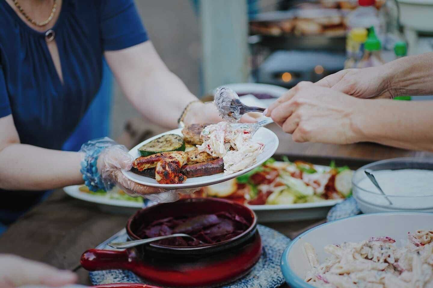 Een groep mensen serveert eten aan een tafel met diverse gerechten, waaronder gegrild vlees en salades.