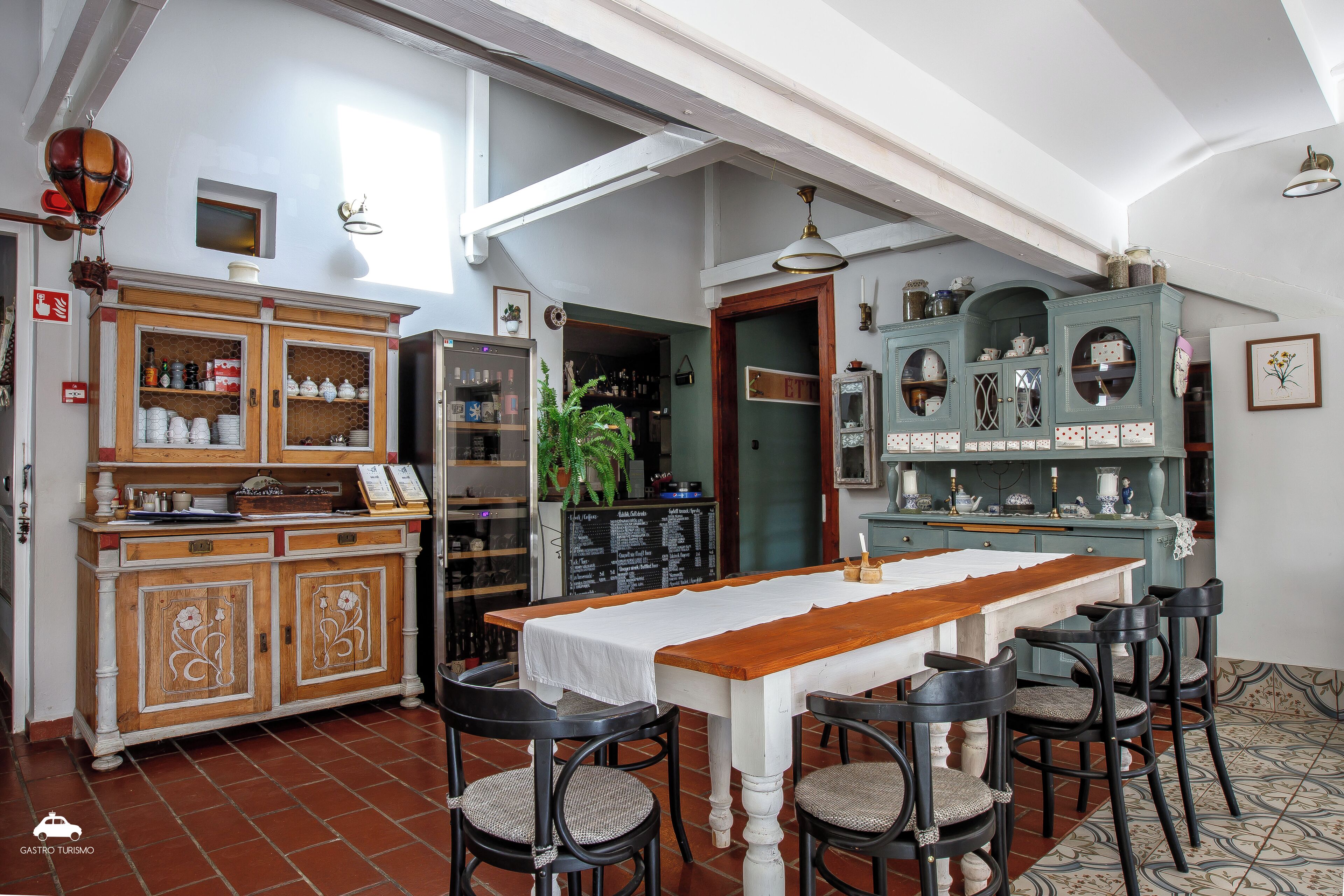 A dining area with a long wooden table, four chairs, and a white tablecloth. There are two wooden cabinets with glass doors, one with a floral design and the other with a geometric pattern. A green plant is visible in the background.