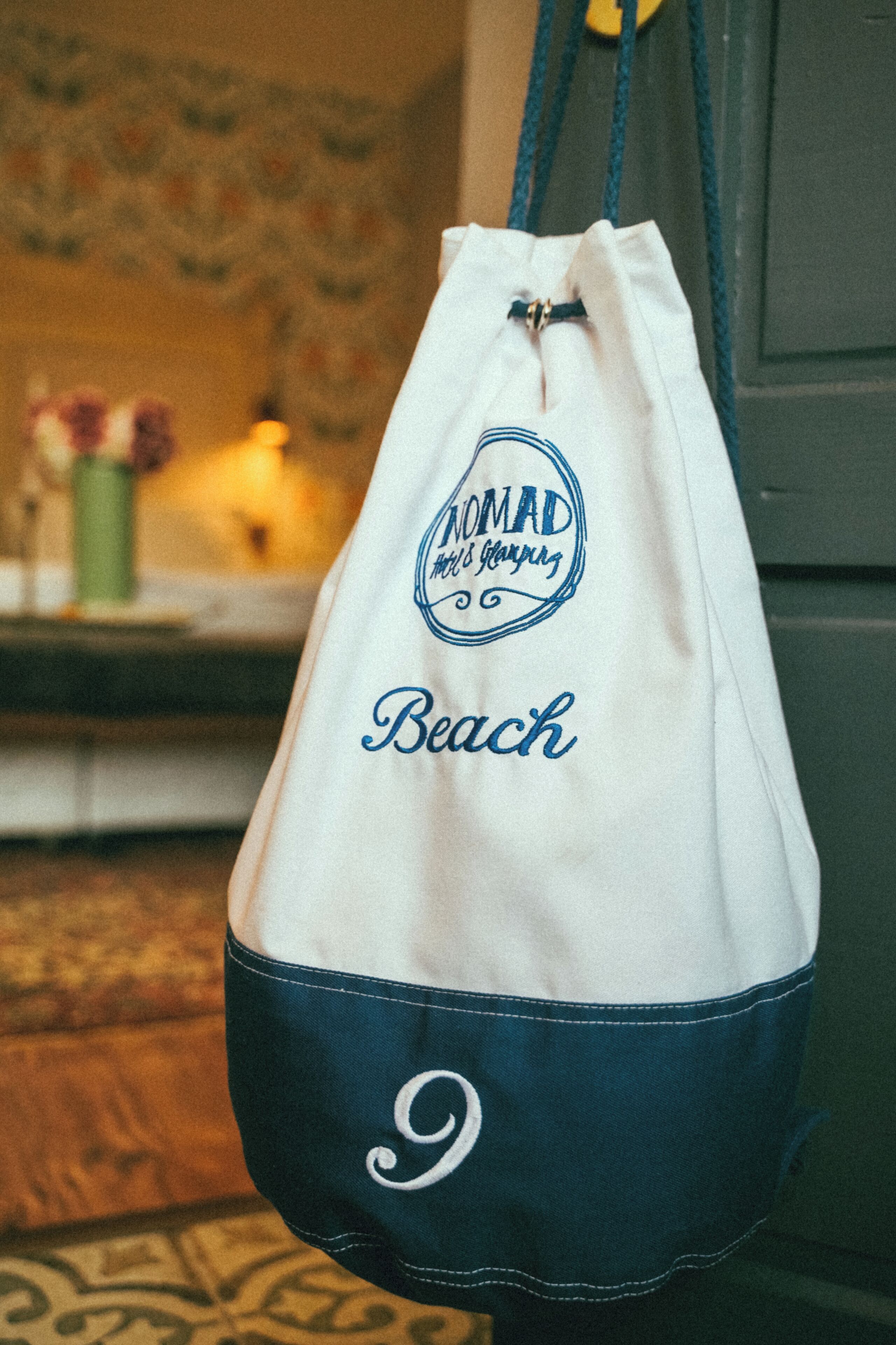 A white and navy blue drawstring bag with the text "Nomad's Calling Beach" and a number "9" on it.