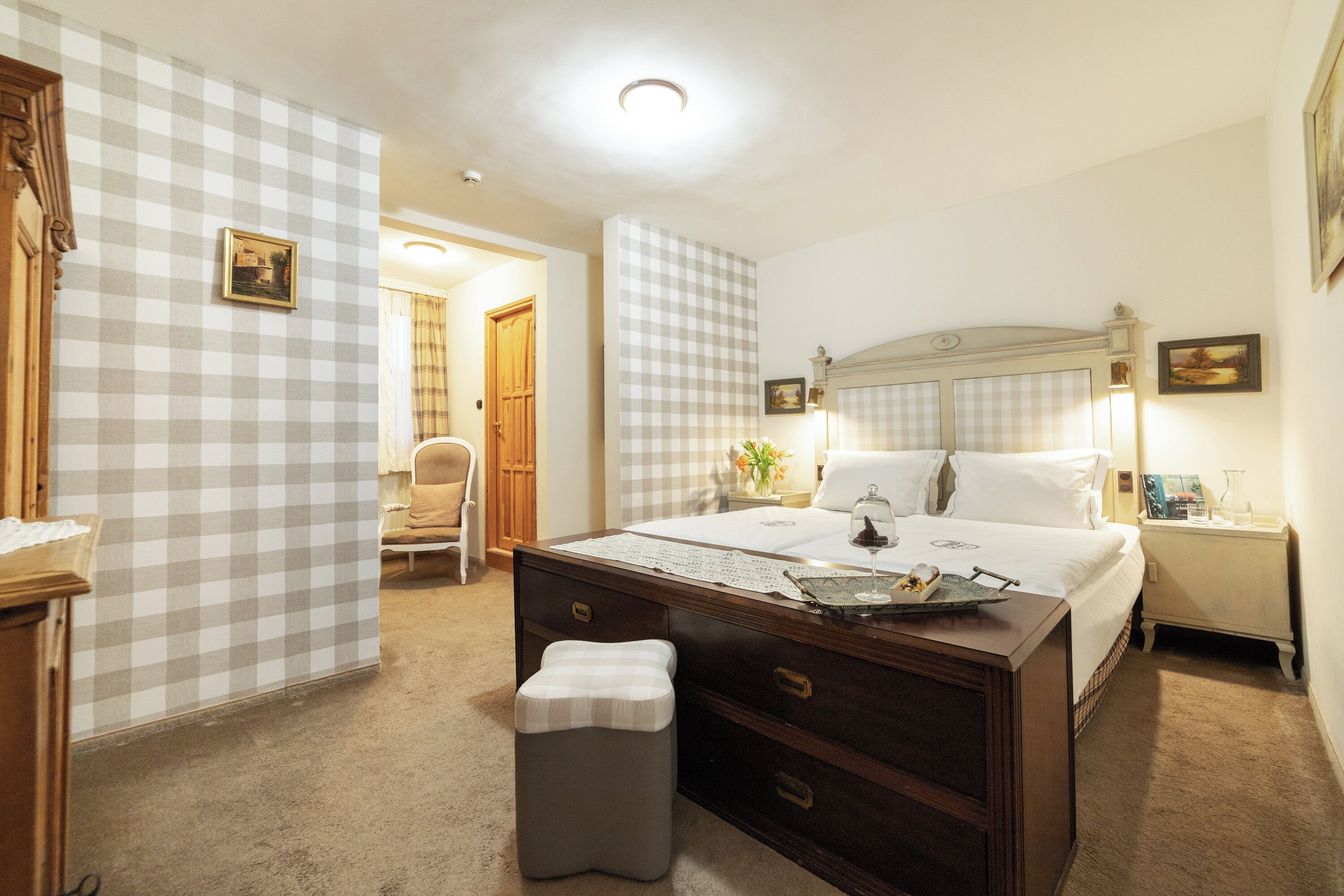 Une chambre d’hôtel avec un grand lit, un bureau, une chaise et un petit tabouret. Un mur à carreaux et une porte donnant sur une autre pièce se trouvent dans la chambre.