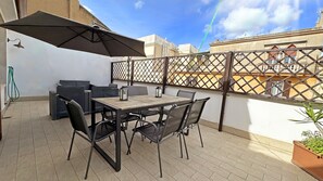 Um pátio externo com mesa e cadeiras, um grande guarda-sol e vista para um edifício ao fundo.