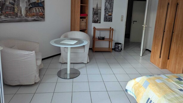 A compact room with a round table, two chairs, a bed, a wooden wardrobe, and a wall clock.