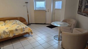 A room with a bed, a chair, a small table, a radiator, and two windows.
