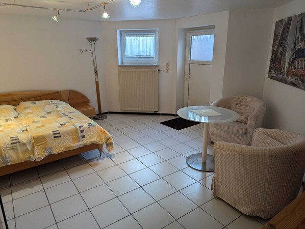 Una habitación con cama, una silla, una mesita pequeña, un radiador y dos ventanas.