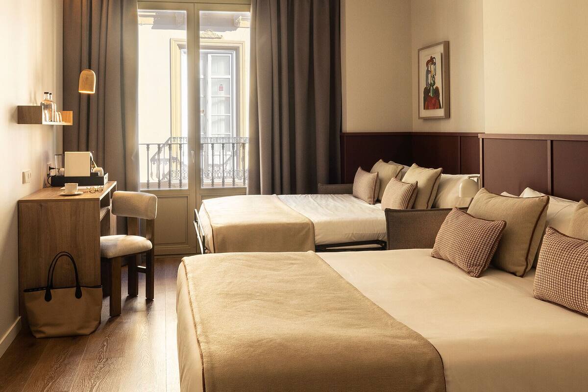 Une chambre d’hôtel avec deux lits, un bureau avec une chaise, un ventilateur de plafond, une fenêtre avec des rideaux et un tableau encadré au mur.