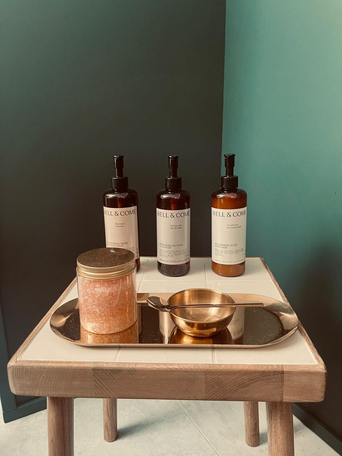 A wooden table with three Well & Come shampoo bottles, a jar of pink salt, and a golden spoon on a tray.