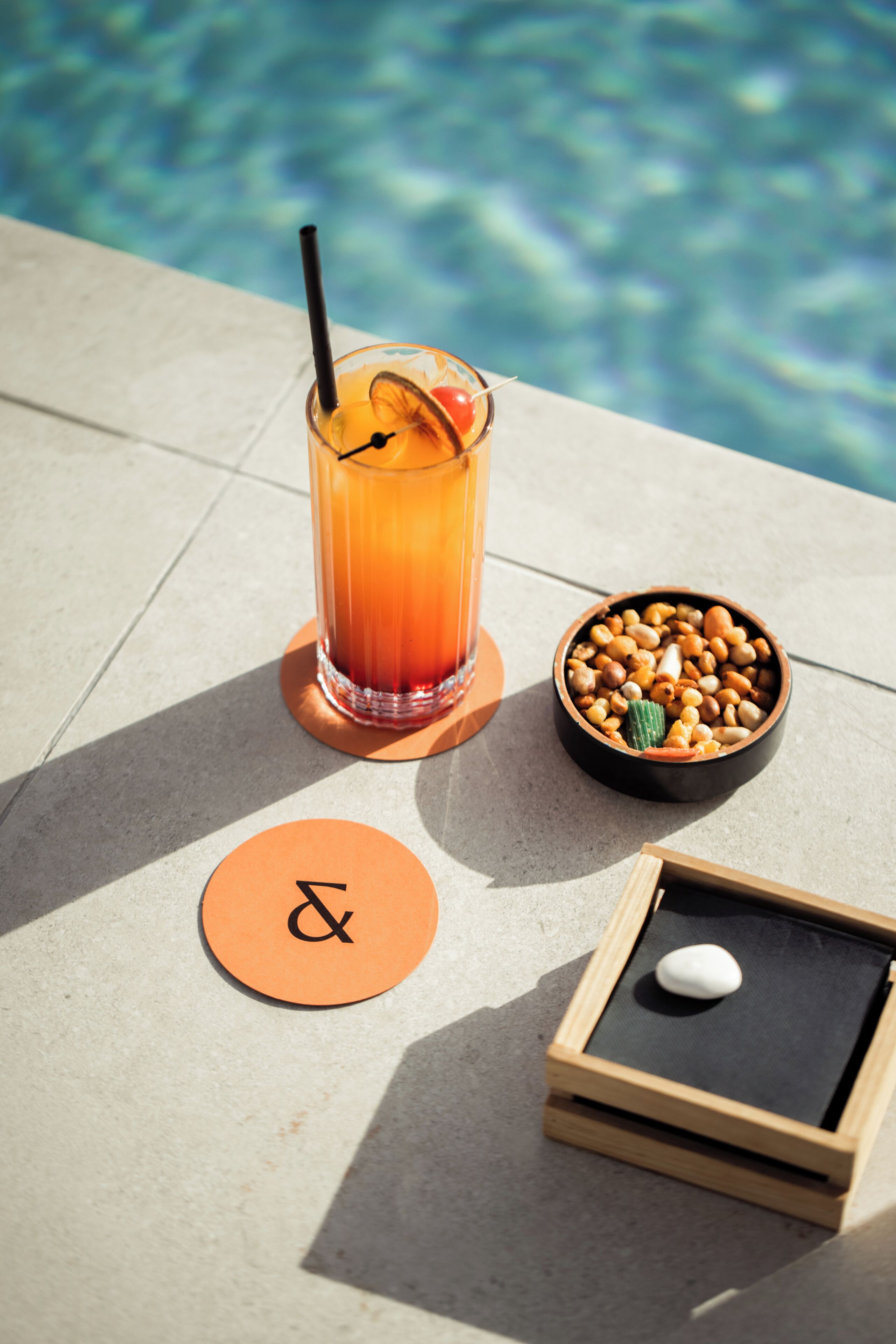 Ein Glas Orangensaft mit Trinkhalm und Orangenscheibe, eine Schale mit Nüssen, eine Holzkiste mit einem weißen Gegenstand und ein runder Orangensaft-Untersetzer mit einem Symbol.