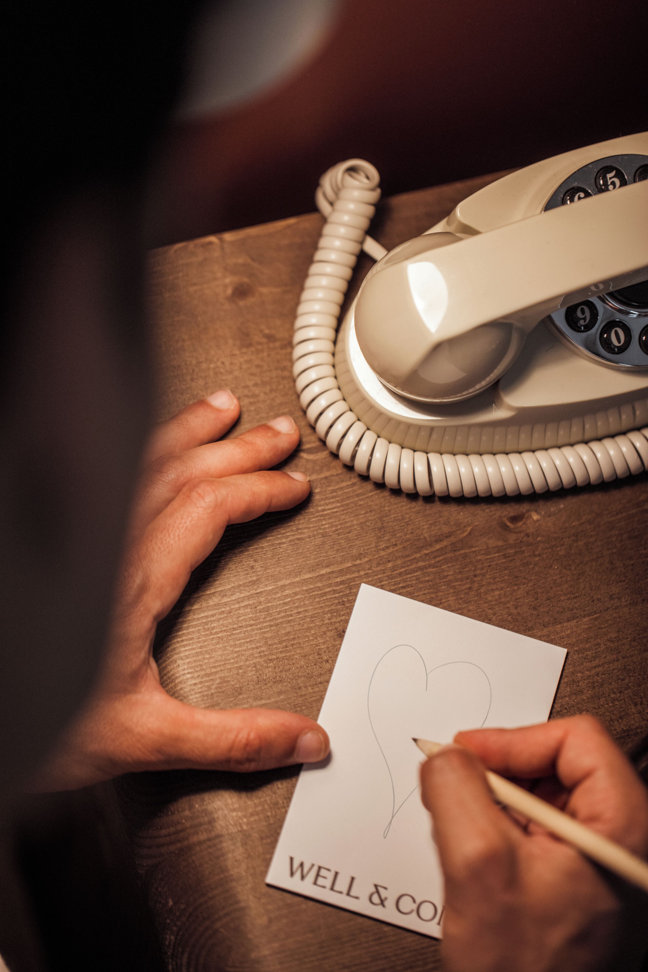 A hand drawing a heart on a piece of paper with the text 'WELL & CO' on it, placed on a wooden surface next to a beige rotary telephone.