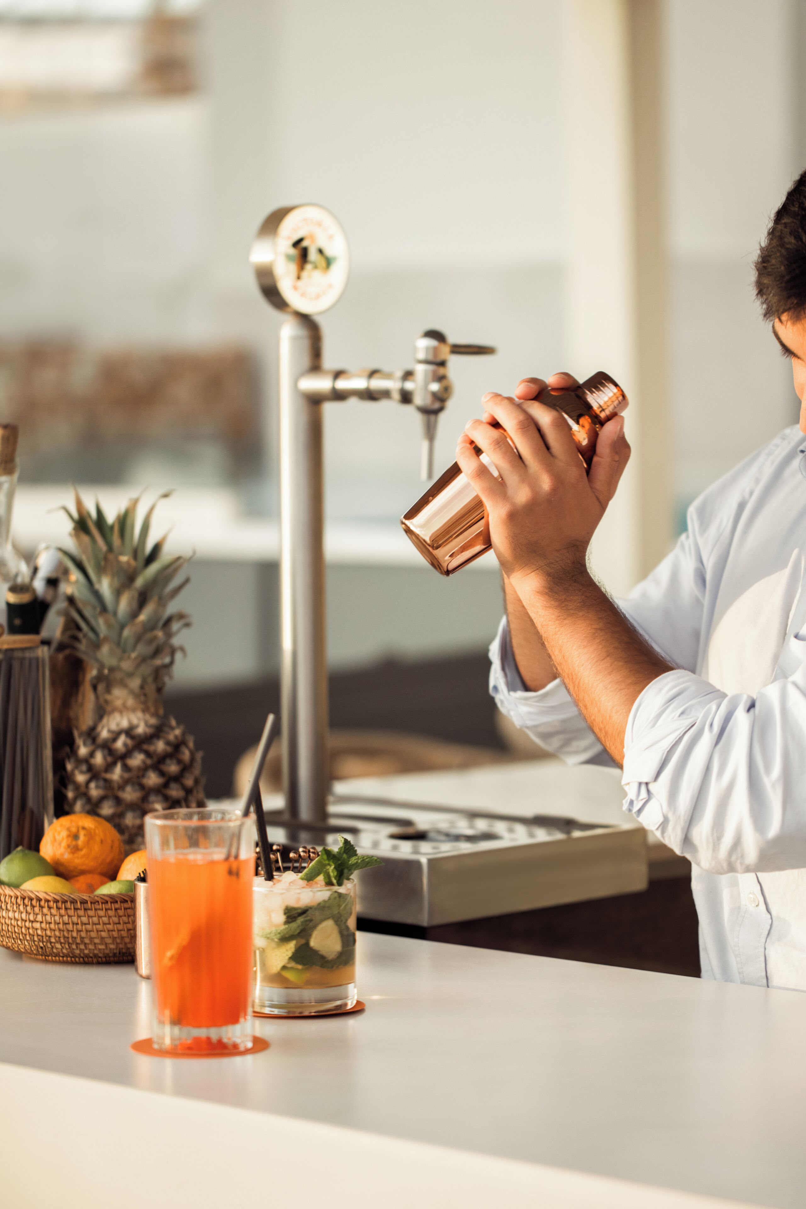 A bartender pouring a drink into a glass, with a pineapple and other fruits on the counter.