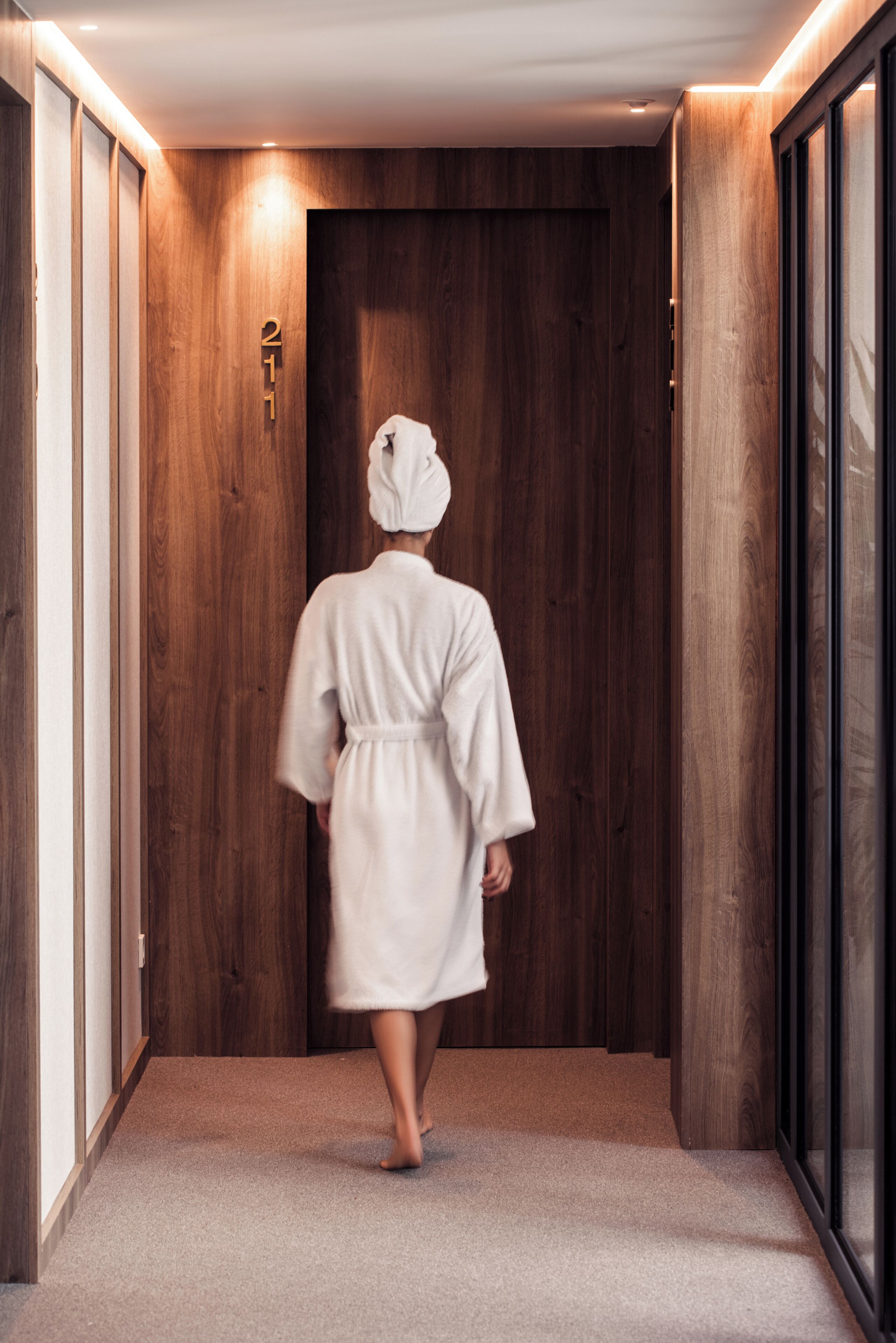 A person in a white robe walking down a hallway with wooden walls and a door marked with the number 21.