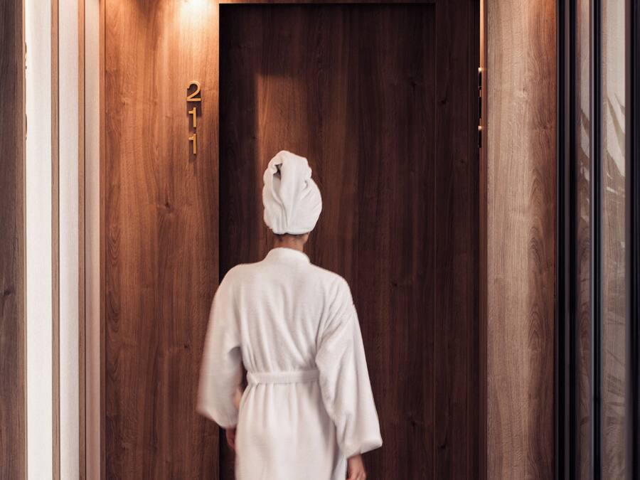 A person in a white robe walking down a hallway with wooden walls and a door marked with the number 21.