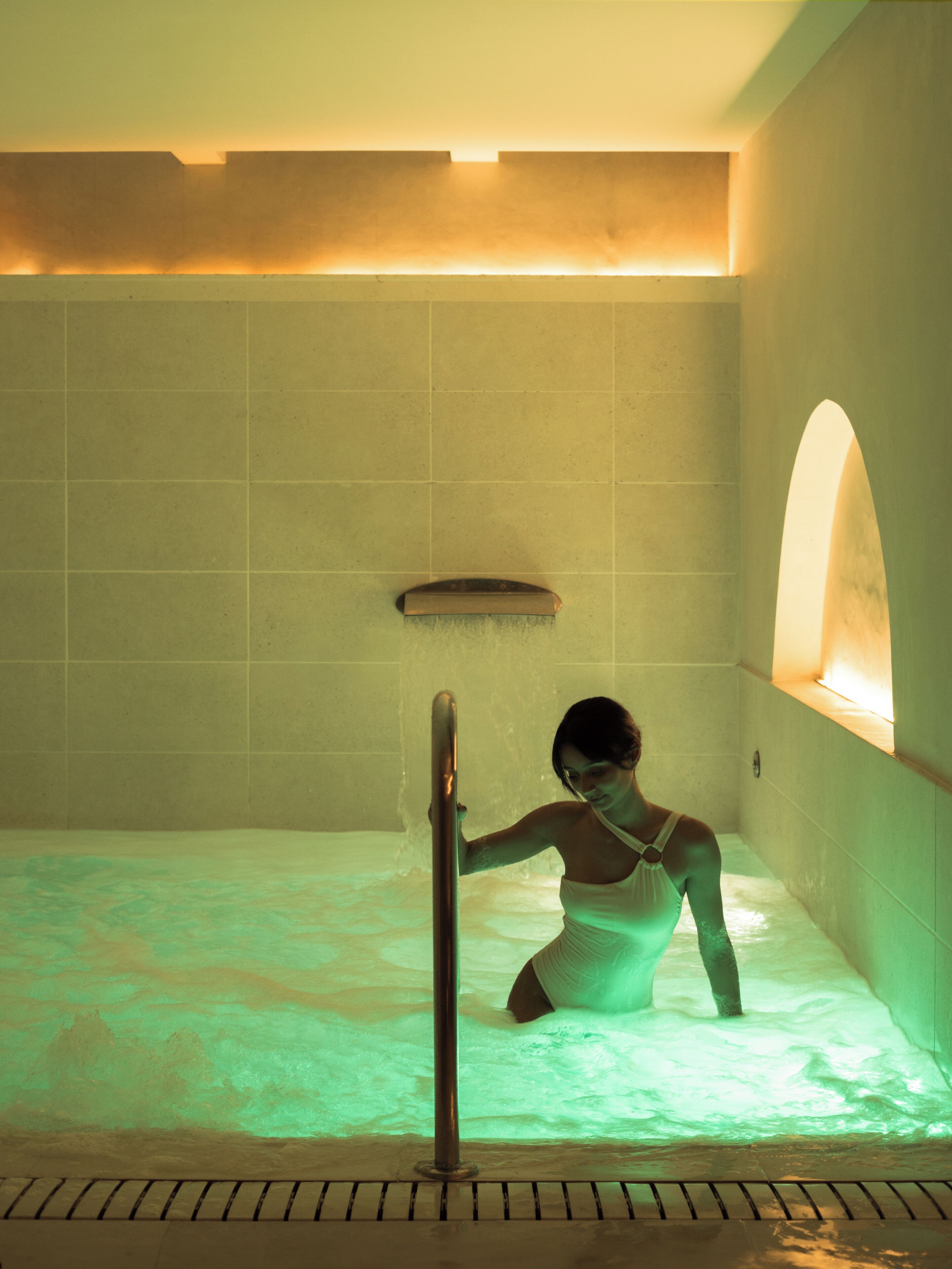 A person in a bathtub with a showerhead, tiled walls, and a bench.