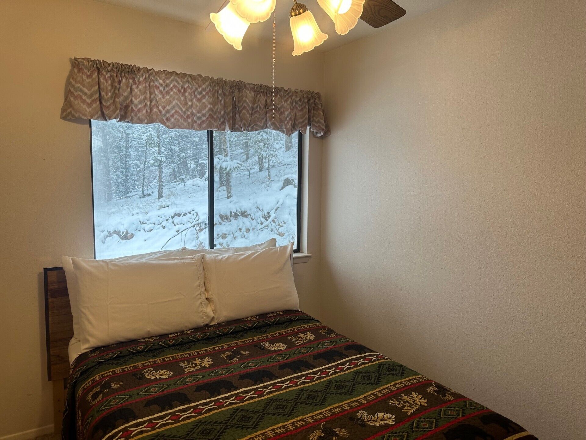 A bedroom with a bed, a window with a view of a snowy forest, a ceiling fan, and a light fixture.