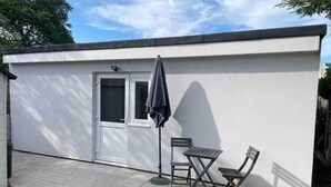 Ein kleines, eingeschossiges Gebäude mit weissem Anstrich, einer Tür und zwei Fenstern. Vor der Tür befindet sich eine Terrasse mit Tisch und Stühlen sowie ein grosser Sonnenschirm.
