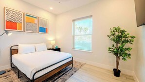 A modern bedroom with a bed, bedside lamps, a window with blinds, and a potted plant. - Coral Cove (South Padre Island)