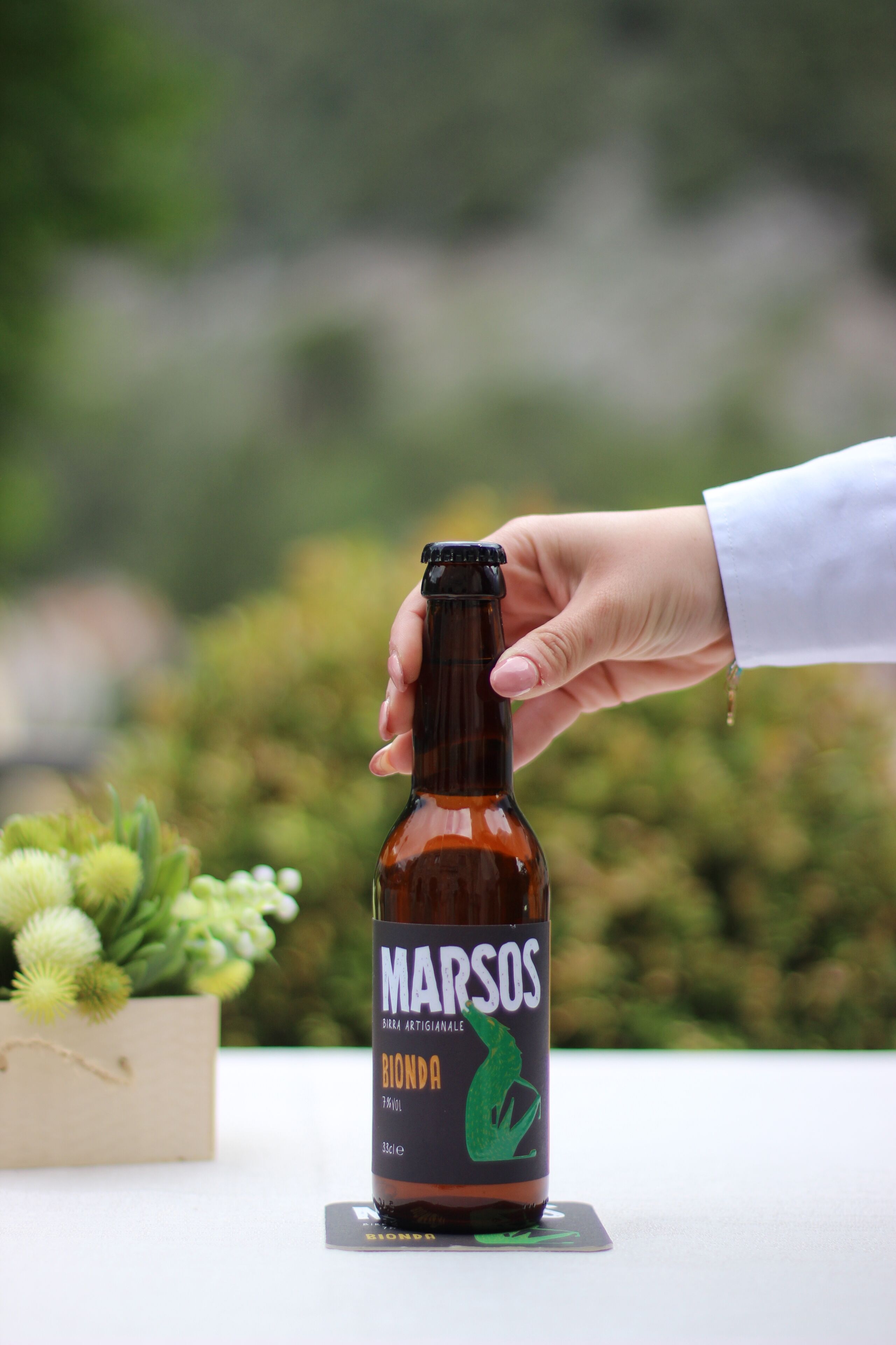 Una mano sostiene una botella de Marsos Blonde Ale con etiqueta que presenta una huella verde.