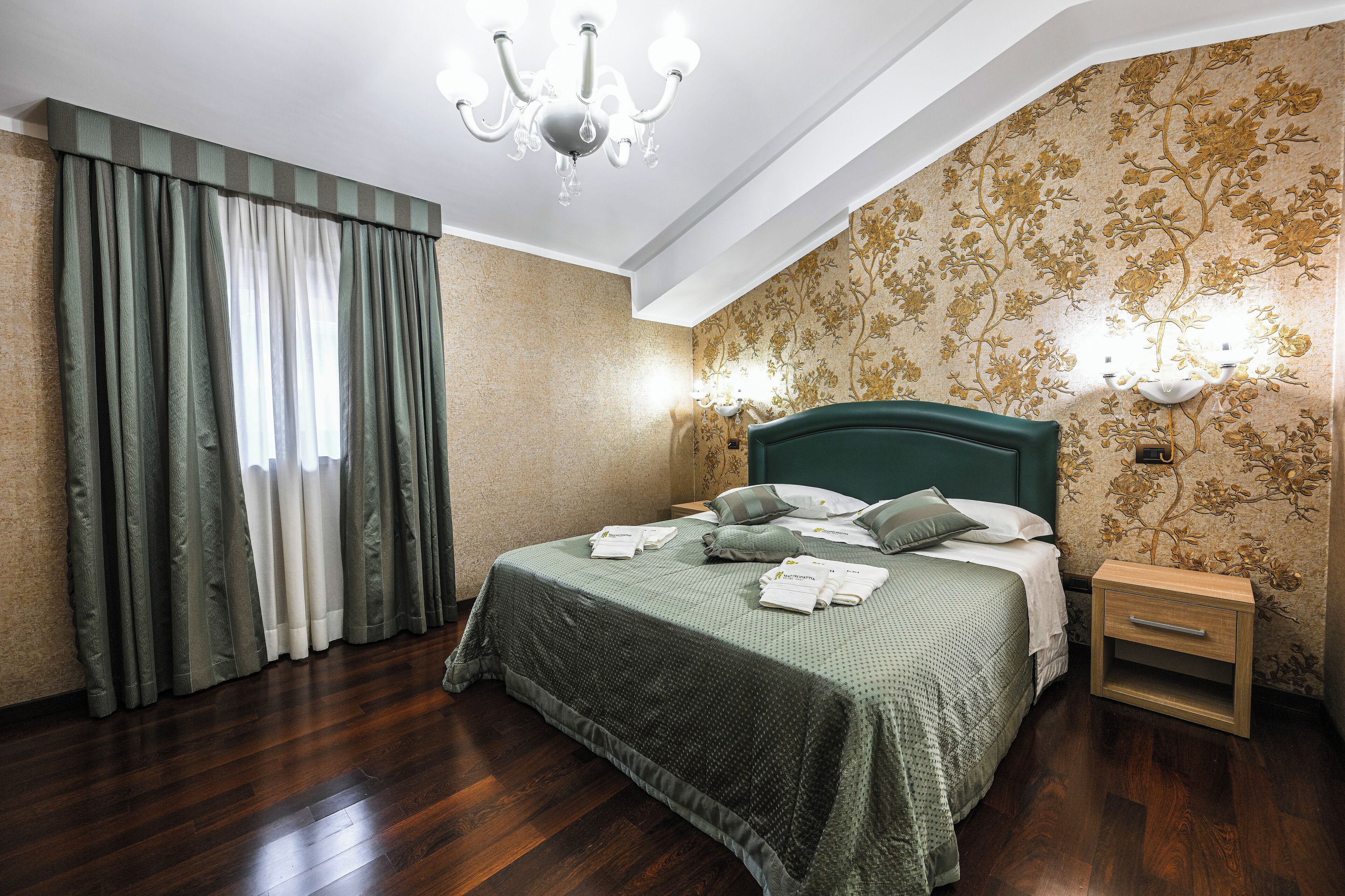 A neatly made bed with pillows and a bedside table, a chandelier, and floral wallpaper.