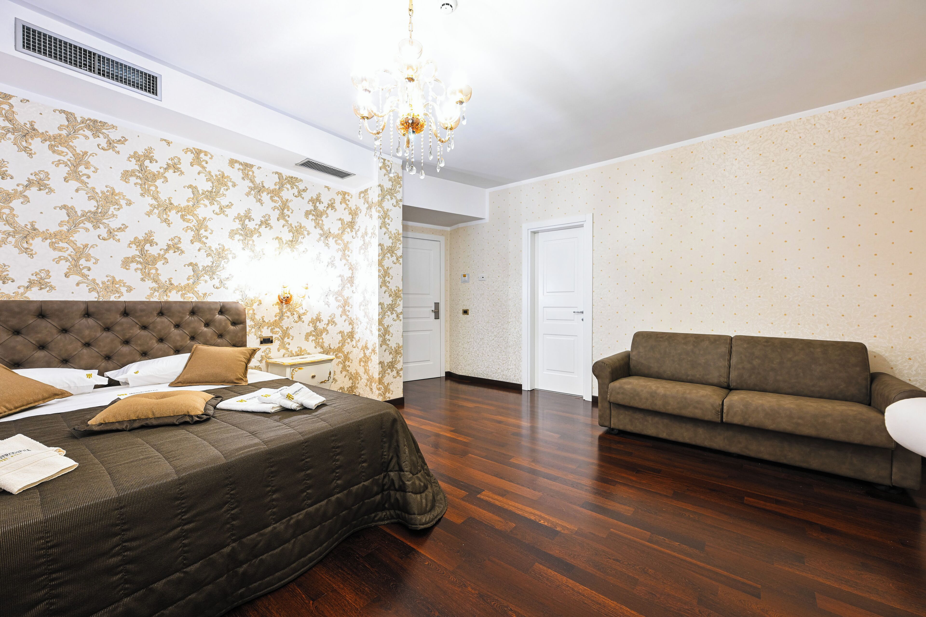 A hotel room with a large bed, a sofa, a chandelier, and patterned wallpaper.