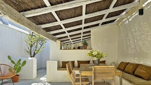 Une terrasse extérieure avec une table en bois et des bancs, entourée de plantes en pot et d’un mur blanc.