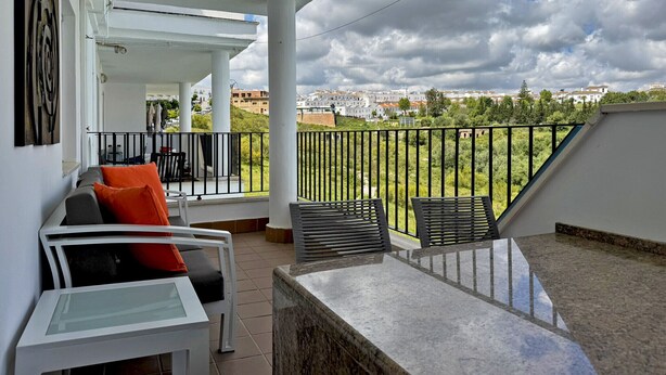 A balcony with a view of a cityscape, a white table, and two chairs with cushions.