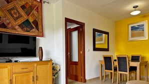 A living room with a wooden cabinet, a glass coffee table with decorative items, a television, and a yellow wall with framed artwork.