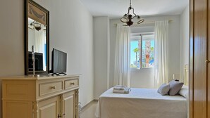 Un dormitorio con cama, televisor sobre una cómoda, ventana con cortinas y vista a palmeras.