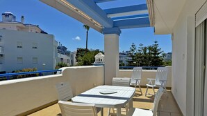 Un balcón con una mesa y sillas blancas, una pérgola y vistas a edificios y árboles.