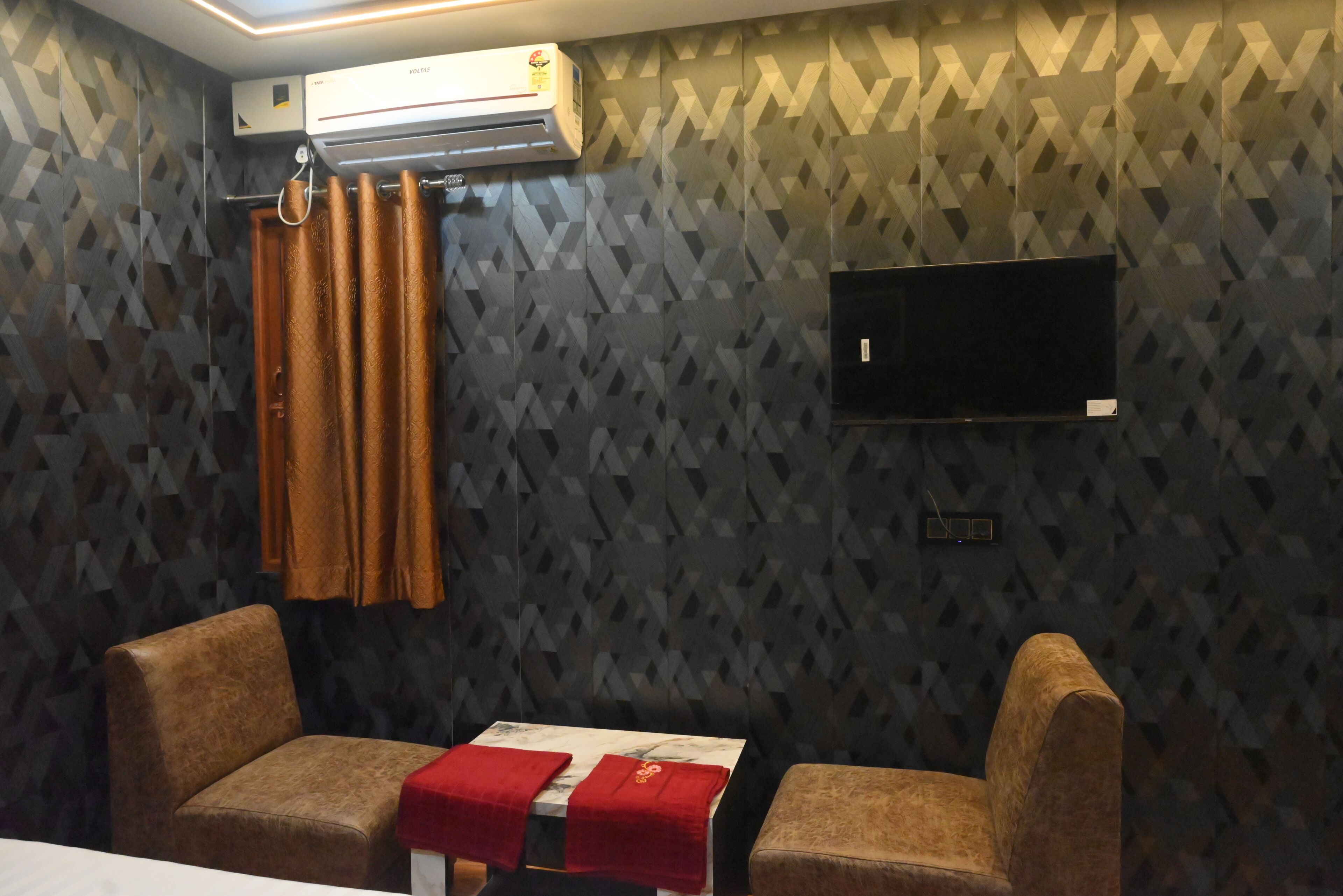 Una stanza con pareti decorate da motivi, una TV a schermo piatto, un tavolo ricoperto da una tovaglia rossa e due poltrone marroni.
