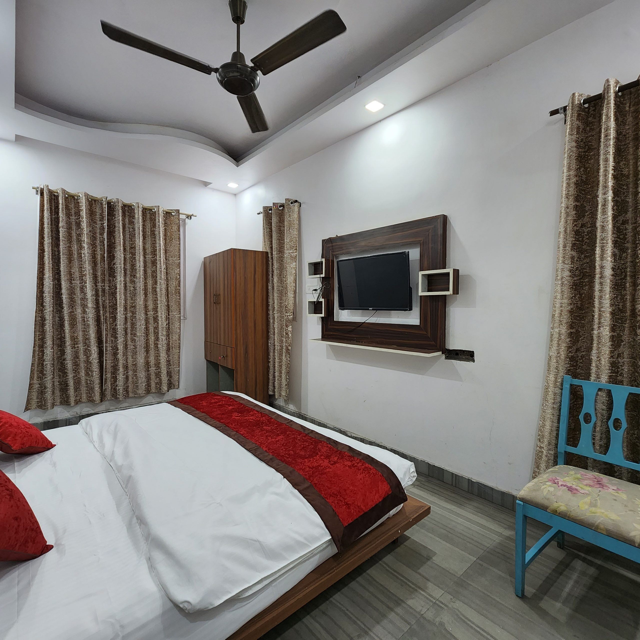 A bedroom with a bed, a TV mounted on the wall, a ceiling fan, and curtains.