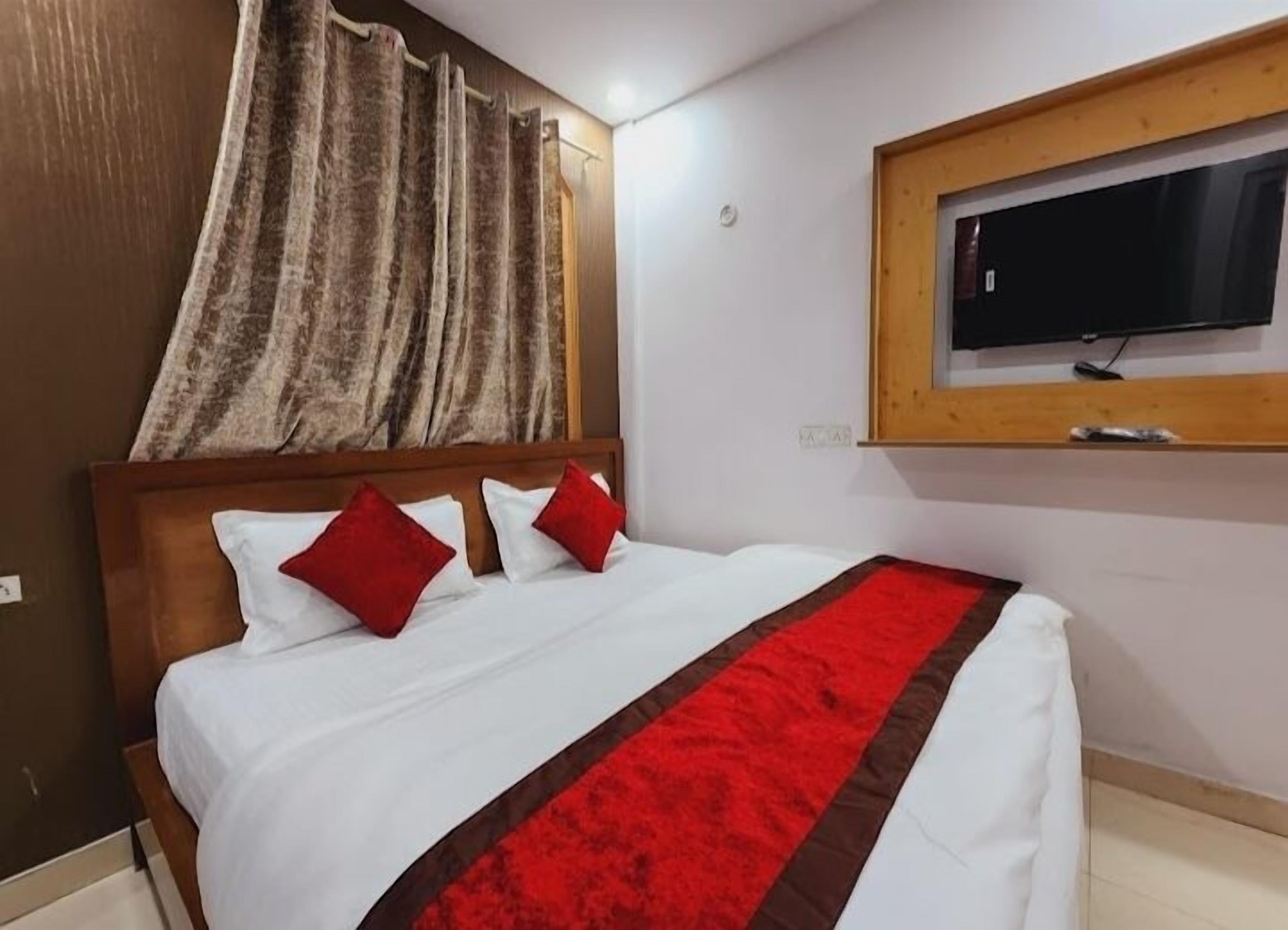 A hotel room with a bed, red and white pillows, a red and brown bedspread, a wooden headboard, a television mounted on the wall, and a window with curtains.