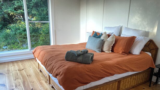 Een groot bed met een houten frame en een bruine sprei, een raam met uitzicht op groen, en een houten vloer.