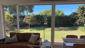 Een woonkamer met een bruine leren bank, een lichtgrijze fauteuil en een houten tafel met stoelen. Grote ramen bieden uitzicht op een gazon en bomen.