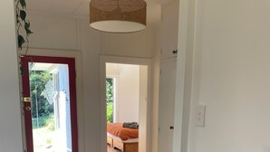 Een hal met houten vloer, een geweven mat, een rode deur en uitzicht op een kamer met een bed en een raam.