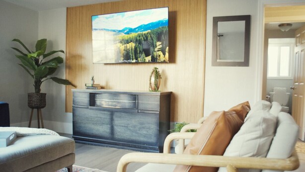 A modern living room with a flat-screen TV mounted on the wall, a grey cabinet with decorative items, a beige sofa with cushions, a potted plant, and a wooden floor. - NEW OSU Reser Stadium Glamour- KING BEDS (Corvallis)