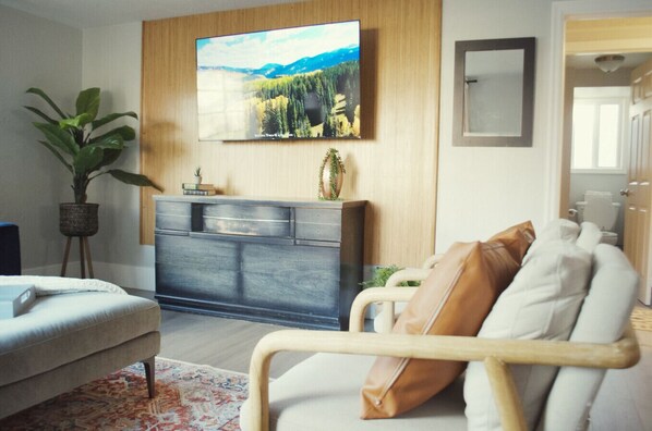 A modern living room with a flat-screen TV mounted on the wall, a grey cabinet with decorative items, a beige sofa with cushions, a potted plant, and a wooden floor.