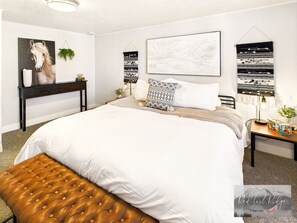 A bedroom with a large bed, a bedside table, a nightstand, a wall-mounted tapestry, and a framed artwork.