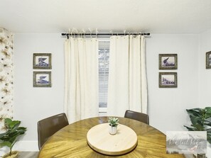 A round wooden table with a central tray, surrounded by chairs, in a room with white walls and curtains.