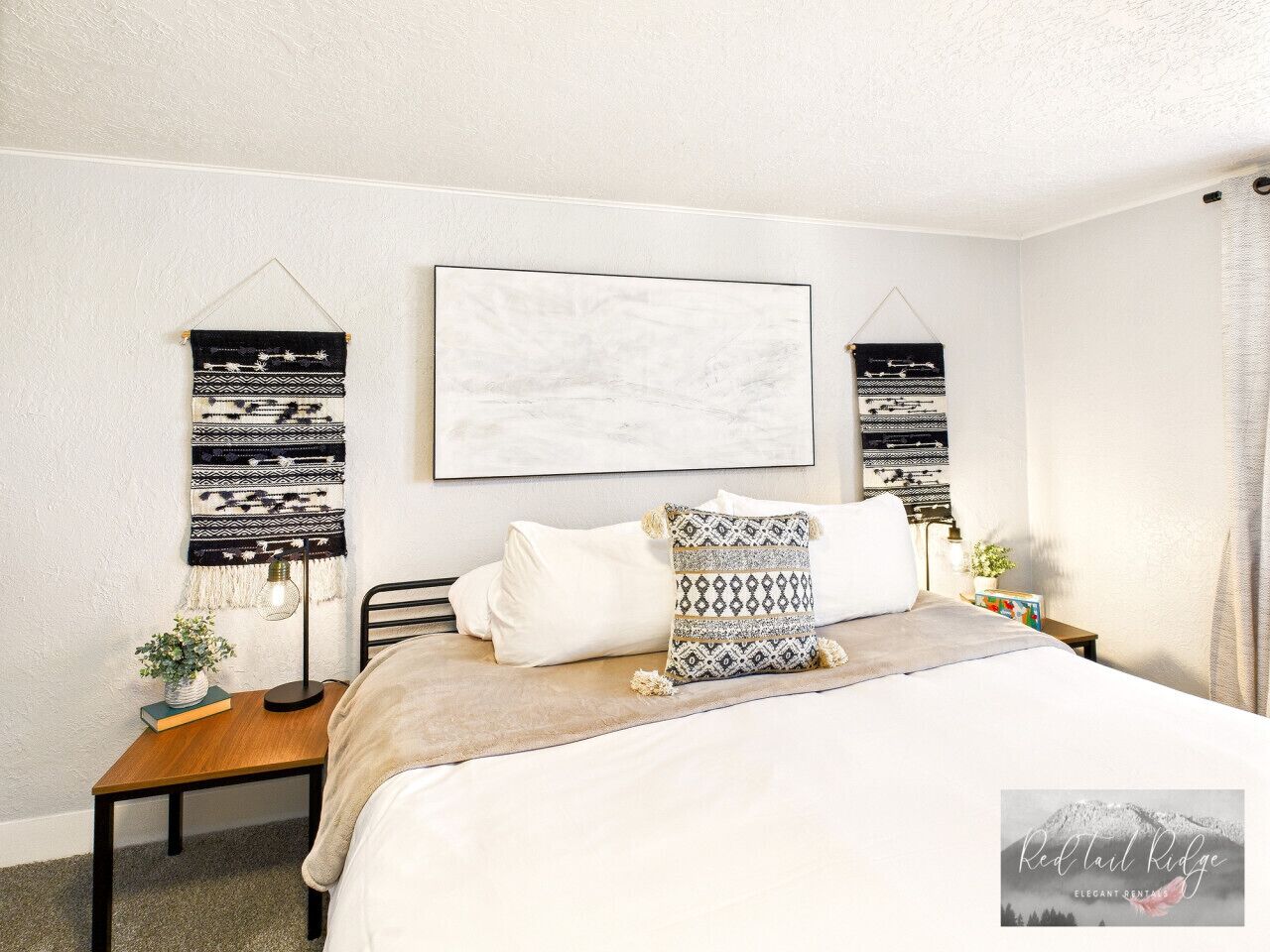 A bedroom with a bed, a bedside table with a lamp, a wall-mounted abstract artwork, and a patterned throw pillow.