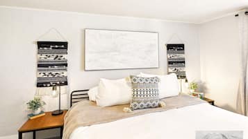 A bedroom with a bed, a bedside table with a lamp, a wall-mounted abstract artwork, and a patterned throw pillow.