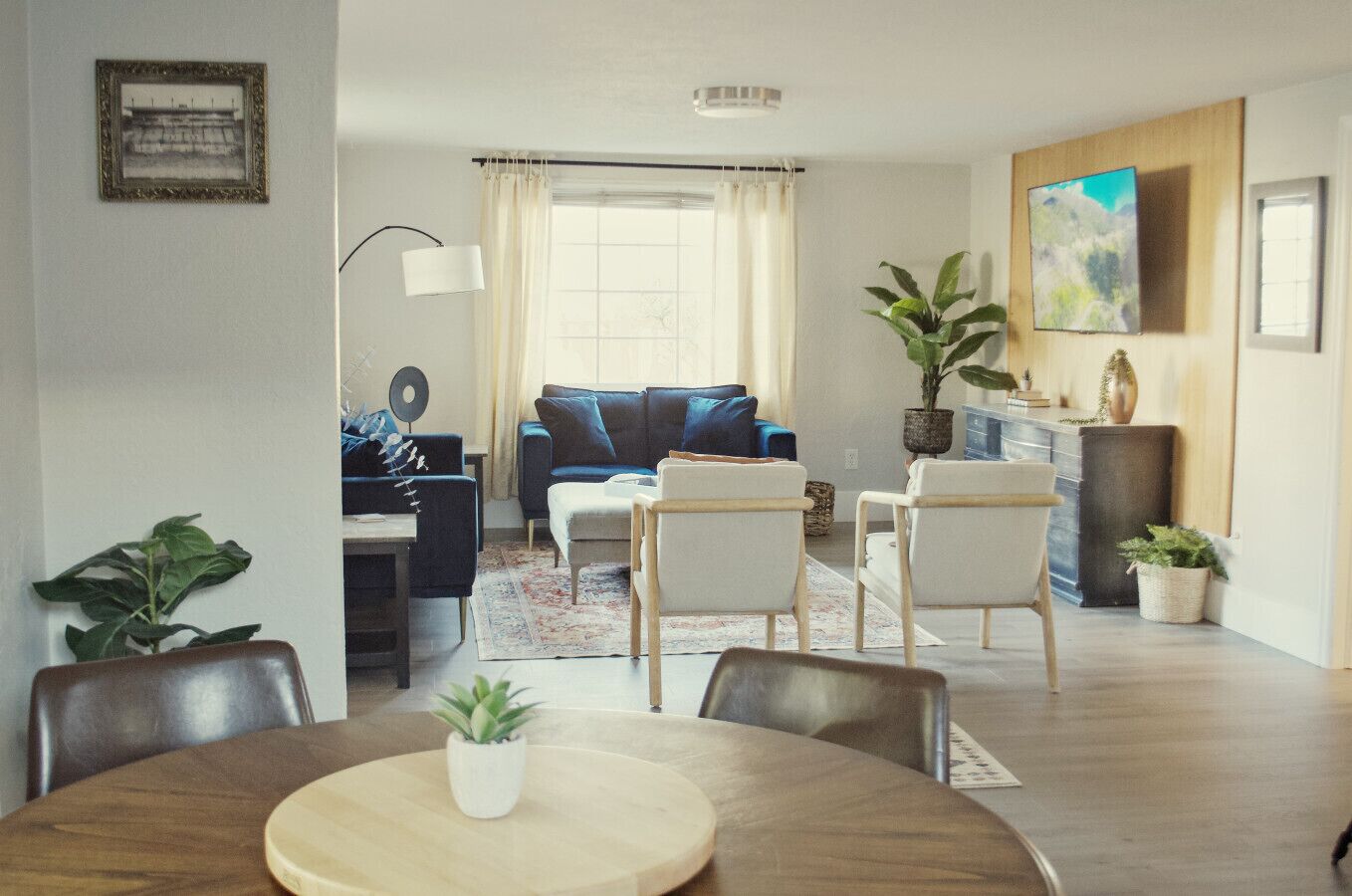A modern living room with a sofa, coffee table, and chairs. There is a dining table with a plant in the center. A TV is mounted on the wall, and a window with curtains is visible.