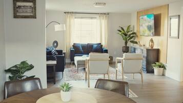 A modern living room with a sofa, coffee table, and chairs. There is a dining table with a plant in the center. A TV is mounted on the wall, and a window with curtains is visible.