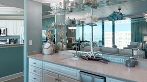 A modern kitchen with a large island, a glass display shelf above the sink, and a "welcome" sign on the countertop.