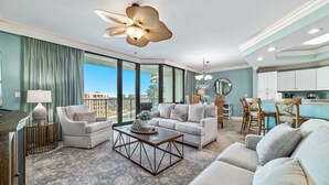 A spacious living room with a large ceiling fan, a coffee table, and a sofa. There is a kitchen area with white cabinets and a dining table with chairs.