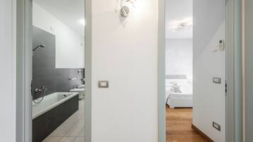 Un bagno moderno con vasca, doccia e servizi igienici. È presente una lampada a parete e una porta che dà su una camera da letto.