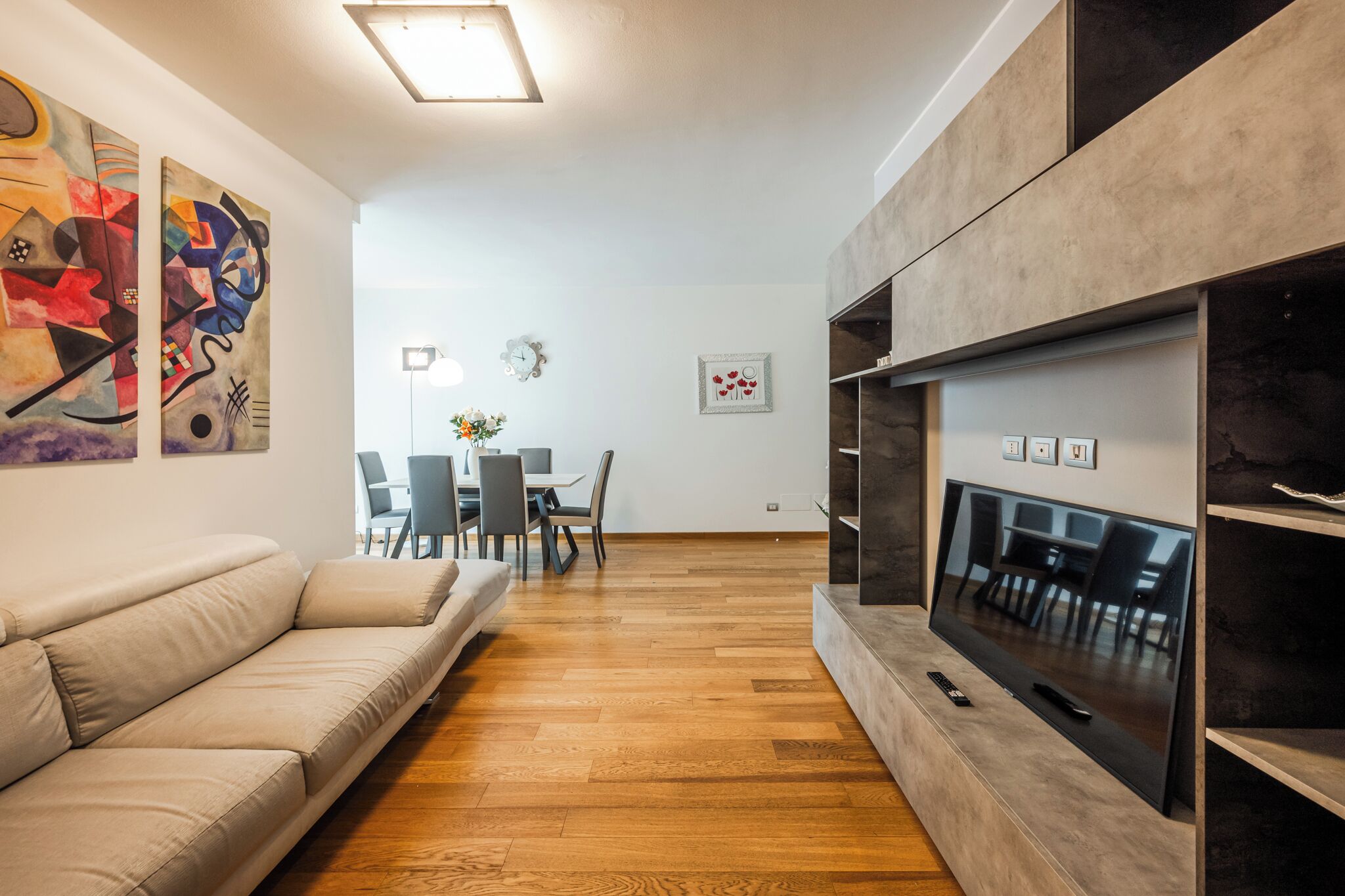 A modern living room with a sofa, a flat-screen TV, wooden flooring, abstract art on the walls, and a dining area in the background.