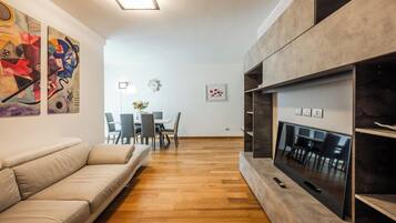A modern living room with a sofa, a flat-screen TV, wooden flooring, abstract art on the walls, and a dining area in the background.