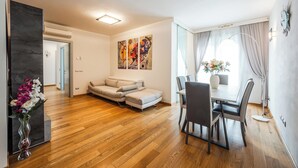 Uma sala de estar moderna com sofá, mesa de jantar e cadeiras. Há um quadro na parede e um vaso com flores no chão.